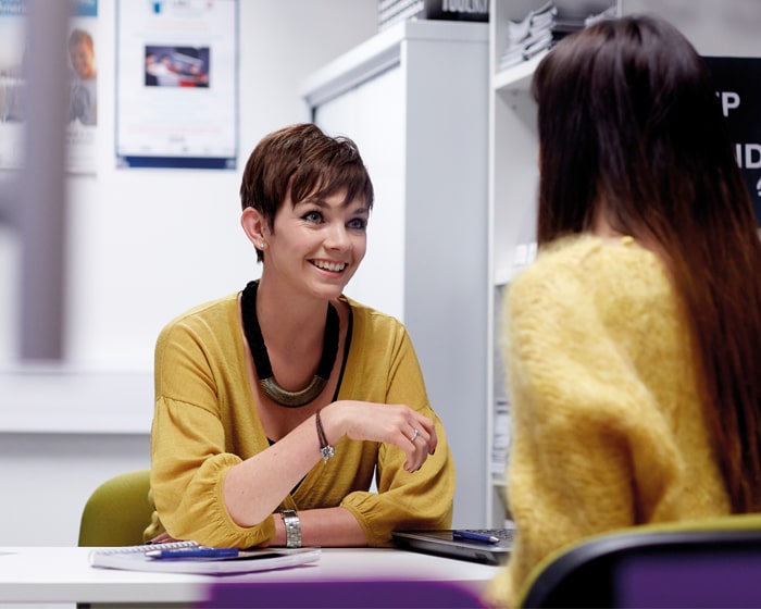 Careers advisor in a yellow top, sat at a table and smiling while she gives advice to s student opposite